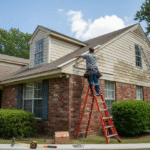 A person on a ladder installs siding on a brick house with tan siding, amidst tools and a sunny sky.